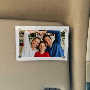 May include: A rectangular photo frame with a family portrait. The frame is attached to a beige surface. The photo shows a woman, a man, and a child smiling and making a heart shape with their hands.