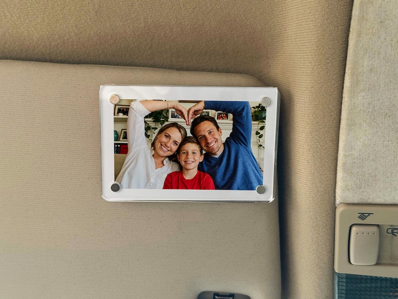 May include: A rectangular photo frame with a family portrait. The frame is attached to a beige surface. The photo shows a woman, a man, and a child smiling and making a heart shape with their hands.