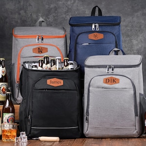 Puede incluir: Cuatro mochilas térmicas en gris, azul marino y negro, cada una con un parche de cuero rectangular con texto personalizado. La mochila negra abierta muestra varias botellas de cerveza de vidrio en su interior, rodeadas de más botellas.
