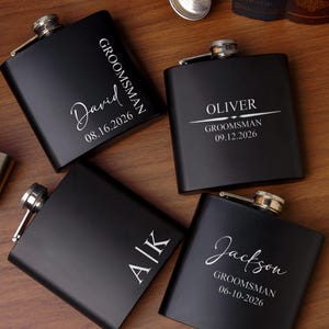 Op de afbeelding: Vier matzwarte metalen heupflacons met zilveren schroefdoppen. Elke flacon is voorzien van aangepaste wit lasergegraveerde tekst, waaronder namen zoals David, Oliver, Jackson, initialen A|K, de titel GROOMSMAN en verschillende data in 2026.