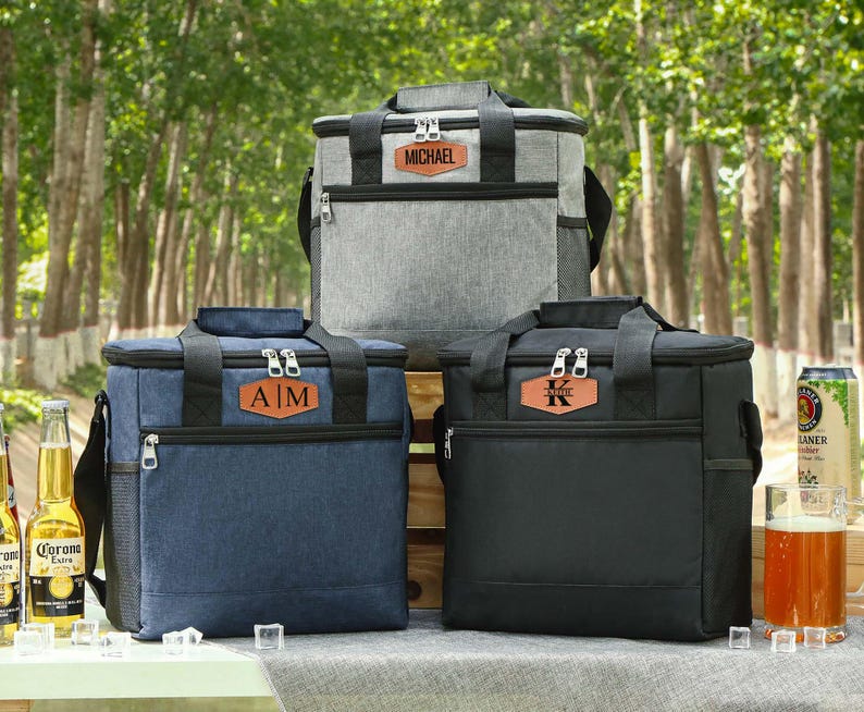 Pu&ograve; includere: Tre borse frigo personalizzate in grigio, blu navy e nero, ciascuna con una toppa in similpelle marrone. Le borse hanno manici e cerniere nere. La borsa grigia ha "MICHAEL" sulla toppa, quella blu "A|M" e quella nera "K".