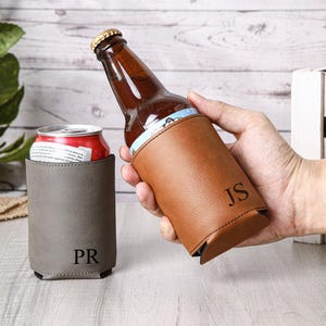 May include: A brown leather beer bottle holder with the initials "JS" and a gray can cooler with the initials "PR". The bottle holder is holding a brown beer bottle. The can cooler is holding a red can.