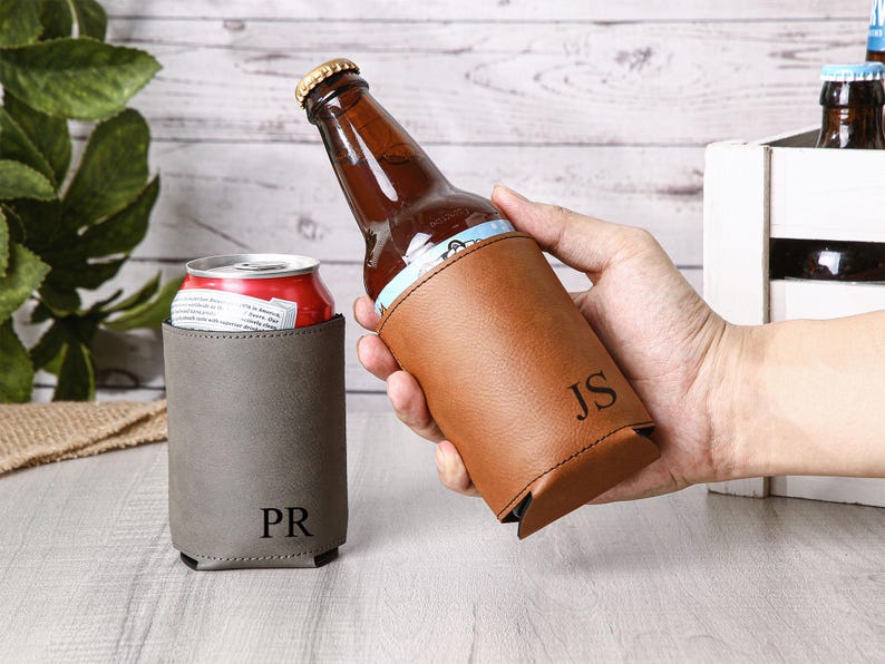 May include: A brown leather beer bottle holder with the initials "JS" and a gray can cooler with the initials "PR". The bottle holder is holding a brown beer bottle. The can cooler is holding a red can.