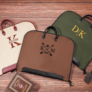 Puede incluir: Tres fundas de ropa personalizadas en crema, marrón y verde oliva, cada una con asa y cierre de cremallera. La bolsa crema tiene "KEVIN" en borgoña, la bolsa marrón tiene un emblema y la bolsa verde oliva "DK" en dorado.