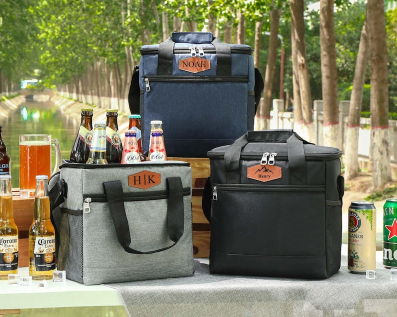 Pu&ograve; includere: Tre borse frigo isolate in blu navy, grigio e nero, ciascuna con un'etichetta personalizzata in similpelle. Le borse sono esposte con bottiglie di birra, un bicchiere di birra e bevande in lattina, suggerendo un picnic o un evento all'aperto.