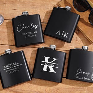 Puede incluir: Cinco petacas de metal negro mate con tapones de rosca plateados, cada una con una personalización única grabada con láser en blanco, incluyendo nombres como Charles, Michael, James, iniciales A|K o KVEIN, fechas y el título GROOMSMAN.