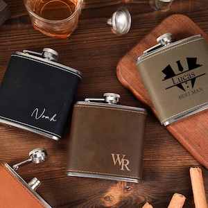 Puede incluir: Tres matraces de cuero marrón con detalles en metal plateado. Los matraces están grabados con los nombres "Noah", "Lucas Best Man" y "WR".