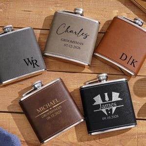 May include: Five personalised hip flasks in grey, tan, brown, and black. Each flask has a silver cap and is engraved with names, initials, and dates. The flasks are arranged on a wooden surface.