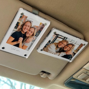 Può includere: Due porta foto trasparenti in acrilico per parasole auto, ognuno con una foto di due donne sorridenti. I supporti sono rettangolari con angoli arrotondati e una clip in metallo. Il testo "Soul sister" è scritto su uno. La visiera è beige.