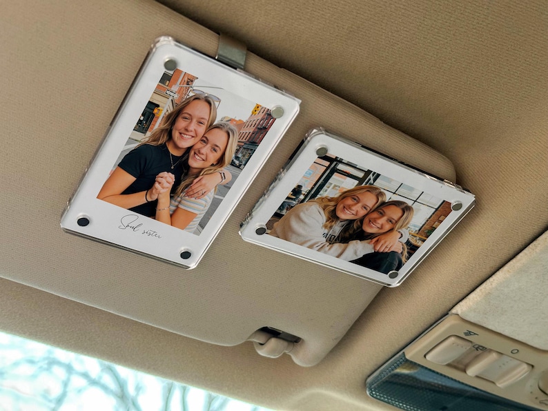 May include: Two clear acrylic car visor photo holders, each displaying a photo of two smiling women. The holders are rectangular with rounded corners and a metal clip. The text "Soul sister" is written on one. The visor is beige.