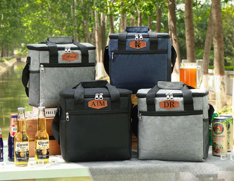 Pu&ograve; includere: Quattro borse frigo personalizzate in grigio, blu navy e nero, con targhette nominative in similpelle marrone. Ogni borsa ha una maniglia superiore, una tracolla e una tasca frontale con cerniera. Le borse sono esposte all'aperto con bevande.