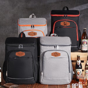 Peut inclure: Plusieurs sacs à dos isothermes noirs et gris, avec des bordures orange et des étiquettes nominatives personnalisées. Des bouteilles de bière et un flacon sont disposés autour des sacs, suggérant un pique-nique.
