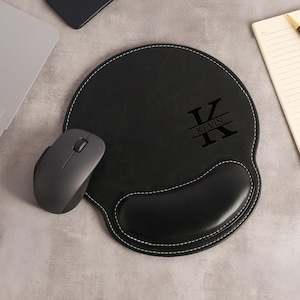 May include: Black leather mouse mat with a wrist rest and white stitching. The mat features a personalized design with the name "KEVIN" and a large initial. A wireless mouse and a laptop are also in the image.