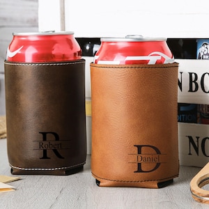 May include: Two personalized leather can coolers, one dark brown and one tan, each holding a red soda can. The dark brown cooler has the name "Robert" and the letter "R" engraved. The tan cooler has the name "Daniel" and the letter "D" engraved.