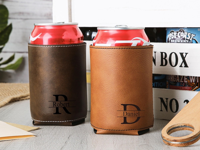 May include: Two personalized leather can coolers, one dark brown and one tan, each holding a red soda can. The dark brown cooler has the name "Robert" and the letter "R" engraved. The tan cooler has the name "Daniel" and the letter "D" engraved.