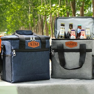 Pu&ograve; includere: Due borse frigo isolate, una blu navy e una grigia, sono affiancate. La borsa blu navy ha le iniziali "A|M" su un'etichetta in pelle, mentre quella grigia ha "JAMES". Entrambe le borse hanno manici e cerniere nere, perfette per picnic e gite.