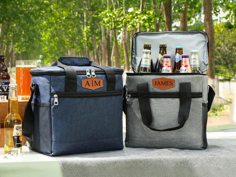 Pu&ograve; includere: Due borse frigo isolate, una blu navy e una grigia, sono affiancate. La borsa blu navy ha le iniziali "A|M" su un'etichetta in pelle, mentre quella grigia ha "JAMES". Entrambe le borse hanno manici e cerniere nere, perfette per picnic e gite.