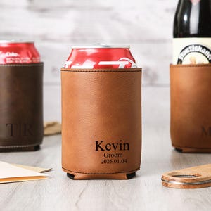 May include: Three leather drink holders, two holding red cans and one holding a bottle. The holders are brown and tan, with personalized text: "TR", "Kevin Groom 2025.01.04", and "Mike". A wooden bottle opener is in the foreground.