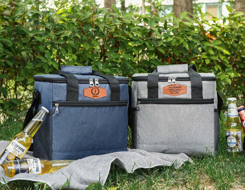 Pu&ograve; includere: Due borse frigo isolate, una blu e una grigia, con dettagli neri e toppe personalizzate in similpelle. La borsa blu ha l'iniziale "J" e il nome "James". La borsa grigia ha il nome "Jackson". Entrambe le borse hanno una maniglia superiore e una tasca frontale con cerniera.