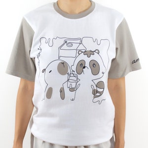May include: White t-shirt with a grey color block on the sleeves. The shirt features a graphic of two cartoon animals, a raccoon and a dog, drinking from a milk carton. The text "am" is on the right sleeve and "ani" is on the left sleeve.