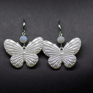 Puede incluir: Un par de pendientes de mariposa plateados. Cada pendiente presenta un colgante de mariposa detallado con patrones de alas grabados y una pequeña cuenta redonda de color azul claro. Los pendientes cuelgan de ganchos plateados sobre un fondo oscuro.