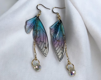 May include: A pair of dangle earrings featuring translucent insect wing shapes in a gradient of purple and teal with gold fleck accents. Each earring hangs from a gold hook and includes a delicate gold chain with a small, clear faceted bead.