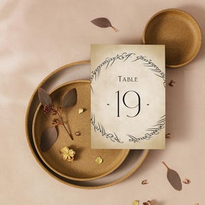 May include: A light brown card with a black circle and the text "TABLE 19" inside the circle. The circle is surrounded by a black script. The card is on a brown plate with a brown background.