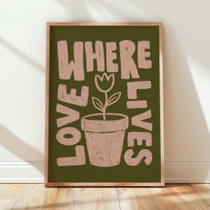 May include: A framed art print with the words "WHERE LOVE LIVES" in a light brown, handwritten style. A light brown flower pot with a flower is in the center. The background is a dark olive green.