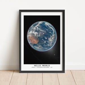 May include: Framed print featuring a photograph of Earth from space, showcasing the planet's blue oceans and brown landmasses against a black background. The text "HELLO, WORLD" is displayed at the bottom, along with the photographer's name and date.