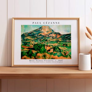 May include: A framed art print featuring Paul Cézanne's painting Mont Sainte-Victoire, c. 1887. The landscape depicts a mountain in shades of blue, green, and orange, with text above and below the image identifying the artist and the work.