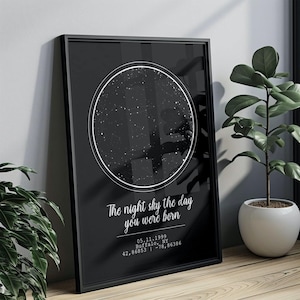 May include: Black framed print featuring a star map within a white circular border. The text "The night sky the day you were born" is displayed below the circle, along with a date, location, and coordinates. A potted plant is in the background.