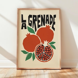 May include: A framed art print on a cream background featuring the text LA GRENADE in bold black letters. The illustration displays two whole orange-red pomegranates and one halved pomegranate showing dark red seeds, accented by green leaves.
