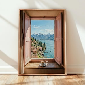 May include: A framed print depicts a scenic view through an open window. The window frame is light brown, with pink shutters. The view shows a lake, mountains, and a village. A cup of coffee and a glass of water sit on the window sill.
