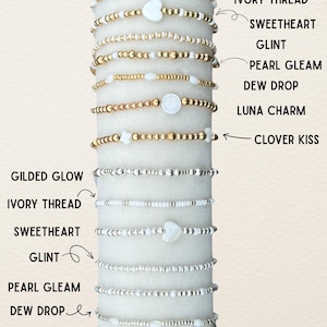 Può includere: Una collezione di braccialetti di perline in tonalità oro e argento, esposti su un cilindro bianco. I braccialetti presentano vari design, tra cui ciondoli a forma di cuore, stella e rotondi. Le etichette di testo identificano ogni stile di braccialetto: Gilded Glow, Ivory Thread, Sweetheart, Glint, Pearl Gleam, Dew Drop, Luna Charm e Clover Kiss.