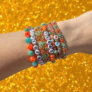 May include: Several beaded bracelets in shades of turquoise, orange, and gold. Some bracelets have white letter beads spelling out "LIFE OF ASH" and "TLOOAS". The bracelets are stacked on a wrist against a gold glitter background.