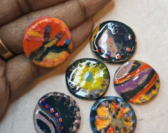 Op de afbeelding: Vijf ronde, platte en glanzende kleiknoppen met handgeschilderde abstracte ontwerpen in levendige kleuren, waaronder oranje, geel, blauw, paars en zwart. Elke knop heeft een gladde, reflecterende afwerking met unieke patronen van stippen, strepen en wervelingen.