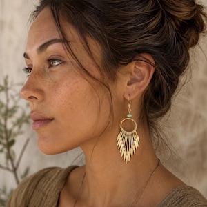 May include: A gold-toned dangle earring featuring a circular hoop frame with a series of thin, pointed, vertical metal fringe elements hanging from the bottom, accented by a small, pale green bead near the top hook attachment point.