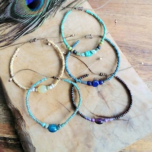 May include: Several handmade beaded bracelets in shades of turquoise, blue, and cream. Each bracelet has a unique bead combination and a sliding knot fastening. The bracelets are displayed on a wooden surface, with a peacock feather in the background.