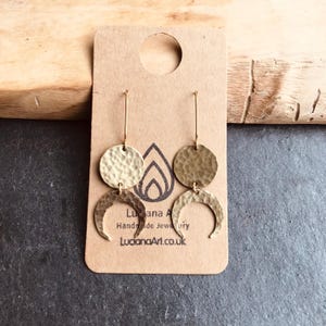 May include: Gold-coloured hammered metal earrings. Each earring has a circular top and a crescent moon shape below. The earrings hang from a thin gold wire. The earrings are displayed on a brown card with the text "Luana A Handmade Jewellery LucianaArt.co.uk".