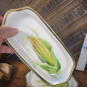 May include: A white porcelain serving dish with a gold rim features a hand-painted image of a corn cob with green husks.