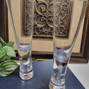 May include: Two clear, stemmed glasses with a flared top and a rounded base. The bases have small bubbles. The glasses are in front of a framed, ornate, gold-colored relief. The background is a dark blue surface.