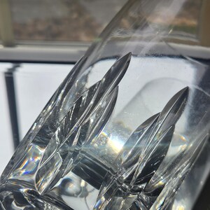 May include: A clear glass tumbler featuring a series of vertical, elongated, leaf-shaped cut patterns around the exterior. The glass has a smooth, polished rim and a thick base, with light refracting through the faceted, geometric design elements.