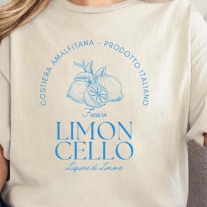 Sand T-shirt with a vibrant blue design reading Costiera Amalfitana - Prodotto Italiano - Limoncello. Minimalist beverage label style. Worn by a woman in jeans, sitting casually. Perfect for limoncello lovers and Italian culture fans.