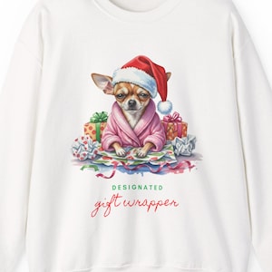 May include: White sweatshirt with a watercolour illustration of a Chihuahua wearing a Santa hat and pink robe, surrounded by wrapped gifts. The text "DESIGNATED gift wrapper" is printed below the image.