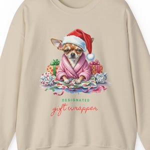Puede incluir: Sudadera beige con un chihuahua con gorro de Papá Noel y bata rosa, rodeado de regalos envueltos y papel. El texto "DESIGNATED gift wrapper" está impreso en escritura roja debajo.