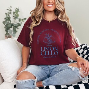 Maroon T-shirt with a vibrant blue design reading Costiera Amalfitana - Prodotto Italiano - Limoncello. Minimalist beverage label style. Worn by a woman in jeans, sitting casually. Perfect for limoncello lovers and Italian culture fans.