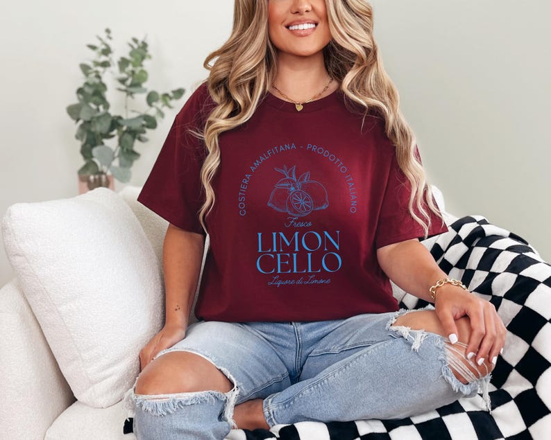 Maroon T-shirt with a vibrant blue design reading Costiera Amalfitana - Prodotto Italiano - Limoncello. Minimalist beverage label style. Worn by a woman in jeans, sitting casually. Perfect for limoncello lovers and Italian culture fans.
