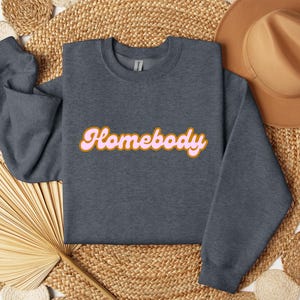 Folded dark heather sweatshirt with a retro cursive design reading Homebody in bold pink with a coffee brown outline. Showcasing the cozy quality of the fabric. Perfect for those who love the comfort of home.