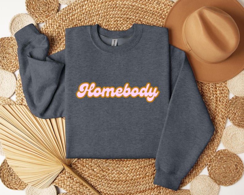 Folded dark heather sweatshirt with a retro cursive design reading Homebody in bold pink with a coffee brown outline. Showcasing the cozy quality of the fabric. Perfect for those who love the comfort of home.