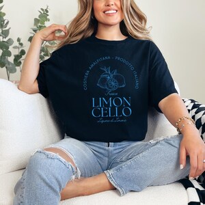 Black T-shirt with a vibrant blue design reading Costiera Amalfitana - Prodotto Italiano - Limoncello. Minimalist beverage label style. Worn by a woman in jeans, sitting casually. Perfect for limoncello lovers and Italian culture fans.